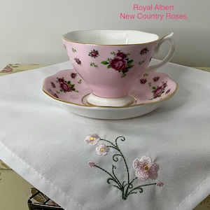 May include: A Royal Albert New Country Roses teacup and saucer set in pink with a floral rose pattern and gold trim, resting on a white cloth napkin featuring an embroidered pink flower design with green stems and leaves on the corner.