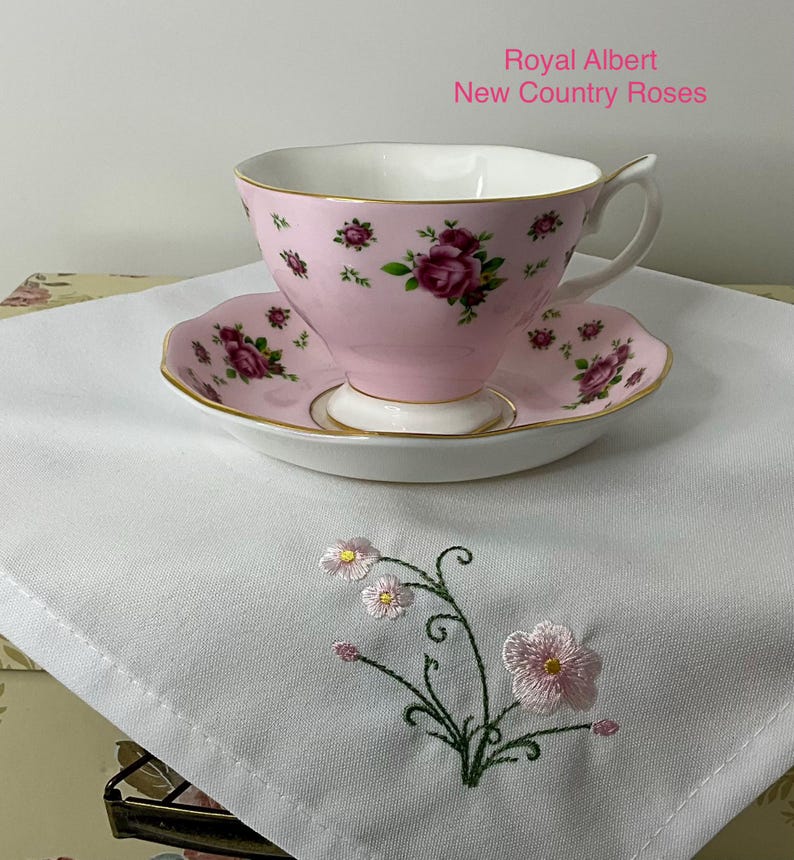 May include: A Royal Albert New Country Roses teacup and saucer set in pink with a floral rose pattern and gold trim, resting on a white cloth napkin featuring an embroidered pink flower design with green stems and leaves on the corner.