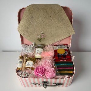 May include: A pink and white striped suitcase gift box containing a tan linen cloth with floral embroidery, a small jar of honey, a candle, two pink rose-shaped soaps, a box of Walkers shortbread, and several tea bags with black and coloured labels.
