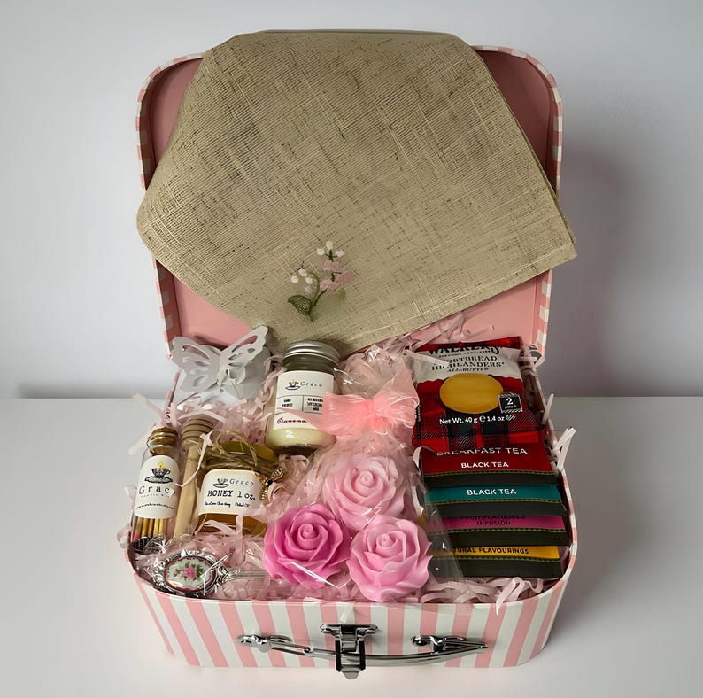 May include: A pink and white striped suitcase gift box containing a tan linen cloth with floral embroidery, a small jar of honey, a candle, two pink rose-shaped soaps, a box of Walkers shortbread, and several tea bags with black and coloured labels.