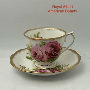 May include: A white porcelain teacup and saucer set featuring the Royal Albert American Beauty pattern. The set is decorated with pink and deep red rose floral motifs, green leaves, and gold trim along the scalloped edges and handle.