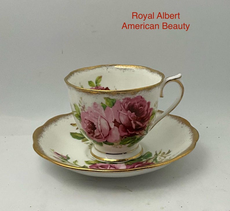 May include: A white porcelain teacup and saucer set featuring the Royal Albert American Beauty pattern. The set is decorated with pink and deep red rose floral motifs, green leaves, and gold trim along the scalloped edges and handle.