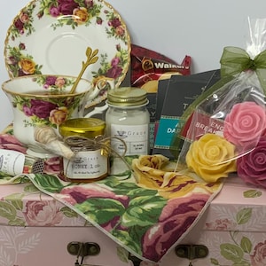 May include: A gift set featuring a floral teacup and saucer, a jar of honey with a wooden dipper, a small soy candle, tea packets, and rose-shaped wax melts. These items are arranged on a pink floral box with a green ribbon and a floral napkin.