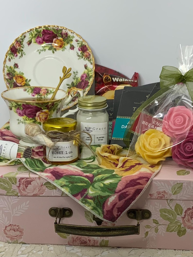 May include: A gift set featuring a floral teacup and saucer, a jar of honey with a wooden dipper, a small soy candle, tea packets, and rose-shaped wax melts. These items are arranged on a pink floral box with a green ribbon and a floral napkin.