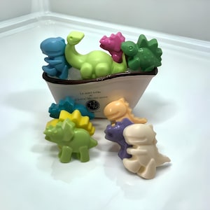 May include: A set of colorful dinosaur-shaped bath toys in a white bathtub. The toys are made of soap and are shaped like different dinosaurs, including a green triceratops, a purple stegosaurus, a blue velociraptor, a green brachiosaurus, and a pink pterodactyl. The bathtub has a French inscription that reads "Le soleil brille et le ciel bleu qui ouvert".