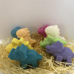 May include: A set of six colorful dinosaur-shaped soaps. The soaps are made in the shape of a triceratops and a stegosaurus. The soaps are in shades of purple, green, yellow, orange, pink, and blue.
