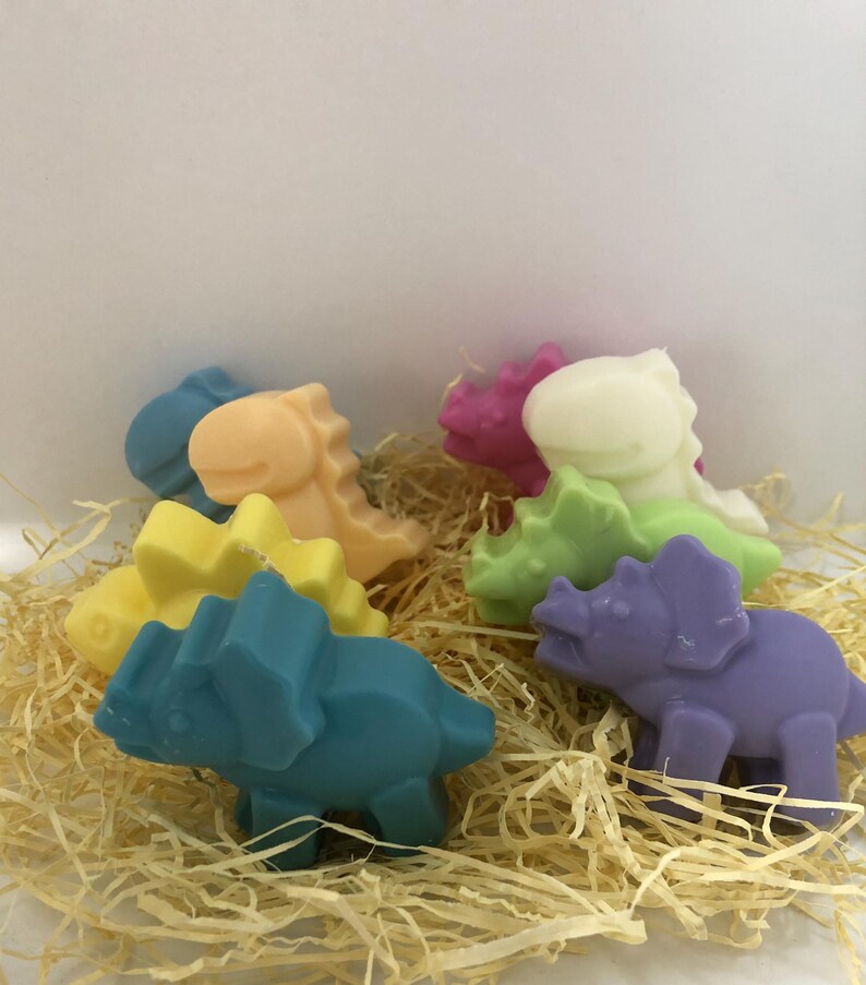 May include: A set of six colorful dinosaur-shaped soaps. The soaps are made in the shape of a triceratops and a stegosaurus. The soaps are in shades of purple, green, yellow, orange, pink, and blue.