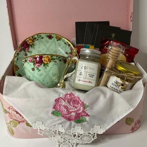 May include: A pink gift box filled with a teacup and saucer, a candle, honey, and a tea towel with an embroidered rose. The box is decorated with floral designs.