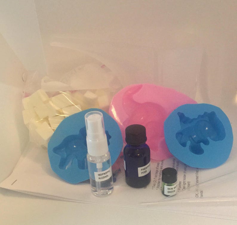 May include: A craft kit for making soap with a variety of supplies, including white soap base, two silicone molds shaped like dinosaurs, a bottle of isopropyl alcohol, a bottle of fragrance oil labeled "Pear", and a small bottle of green soap color.