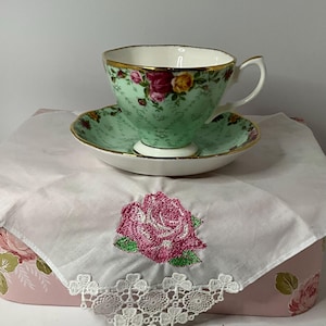 May include: A light green porcelain teacup and saucer set featuring a gold rim and a floral pattern of pink and yellow roses. The set rests on a white fabric napkin with a pink embroidered rose and white lace trim, placed atop a pink floral box.