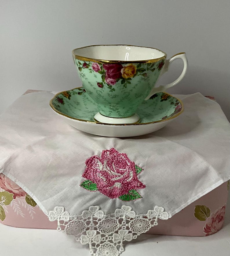 May include: A light green porcelain teacup and saucer set featuring a gold rim and a floral pattern of pink and yellow roses. The set rests on a white fabric napkin with a pink embroidered rose and white lace trim, placed atop a pink floral box.