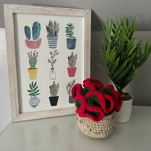 May include: A framed print of a white background with a grid of 12 colorful illustrations of potted plants. The print is hanging on a white wall next to a crocheted red and green plant in a beige pot.