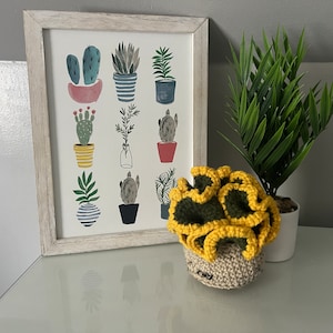 May include: A crocheted succulent plant with yellow and green leaves in a beige pot. The plant is sitting on a white surface next to a framed print of potted plants.