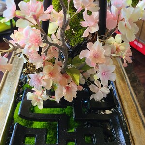 May include: A decorative floral arrangement featuring pink and white cherry blossoms. The arrangement includes green moss, a black geometric design, and a wooden frame. The blossoms are in full bloom, creating a delicate and natural aesthetic.