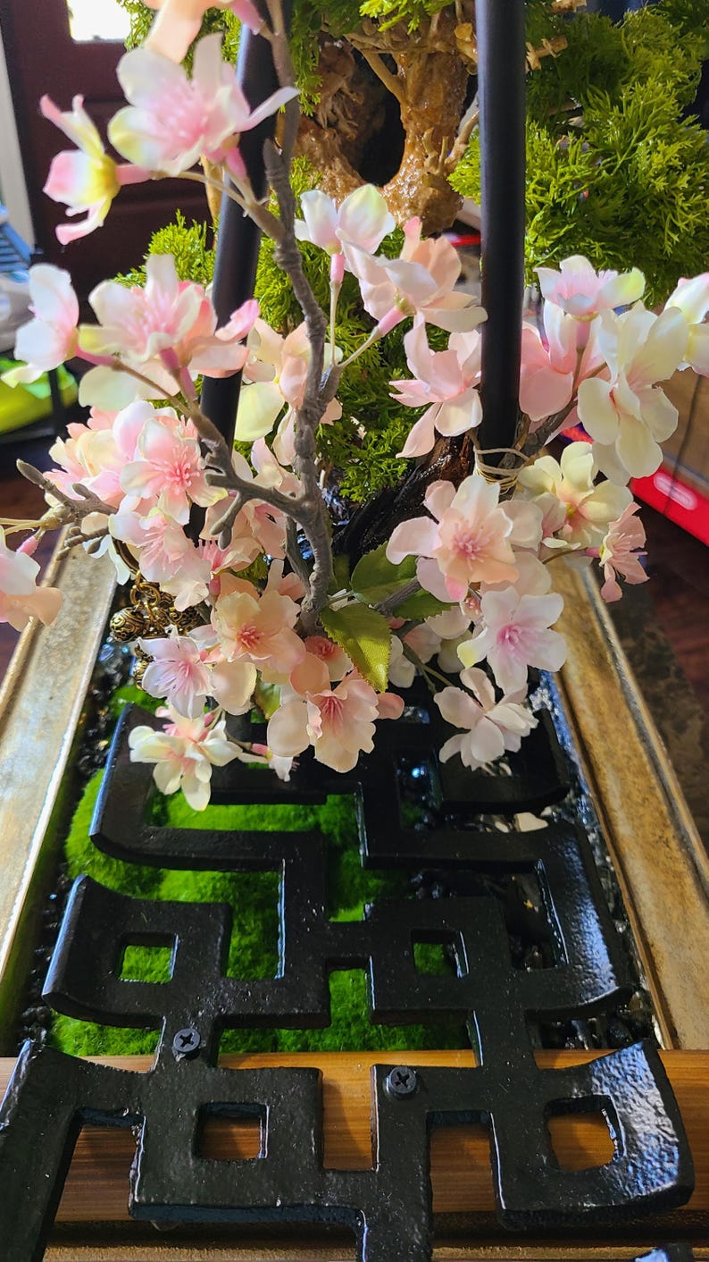 May include: A decorative floral arrangement featuring pink and white cherry blossoms. The arrangement includes green moss, a black geometric design, and a wooden frame. The blossoms are in full bloom, creating a delicate and natural aesthetic.