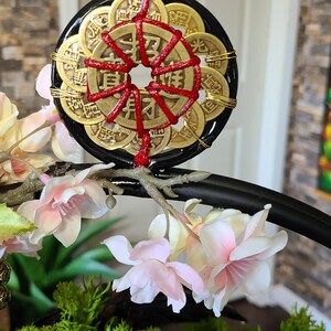 May include: A decorative black and gold coin charm with red accents. The charm is surrounded by gold coins and red string. The charm is displayed with pink and white flowers and green moss.