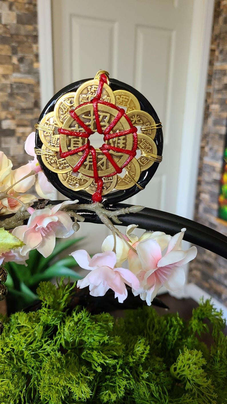 May include: A decorative black and gold coin charm with red accents. The charm is surrounded by gold coins and red string. The charm is displayed with pink and white flowers and green moss.