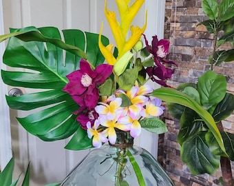 May include: A green glass vase with a round body and a wide mouth, filled with a tropical flower arrangement. The arrangement includes yellow ginger lilies, pink and white plumeria flowers, and purple clematis flowers. A large green monstera leaf is also included in the arrangement.
