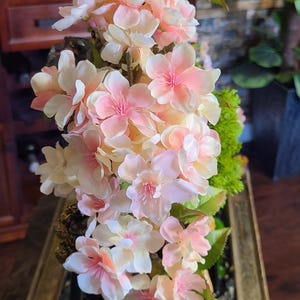 May include: A floral arrangement featuring delicate pink and white blossoms with green leaves. The flowers are arranged in a rectangular, gold-toned container with a dark green interior, creating a decorative display.
