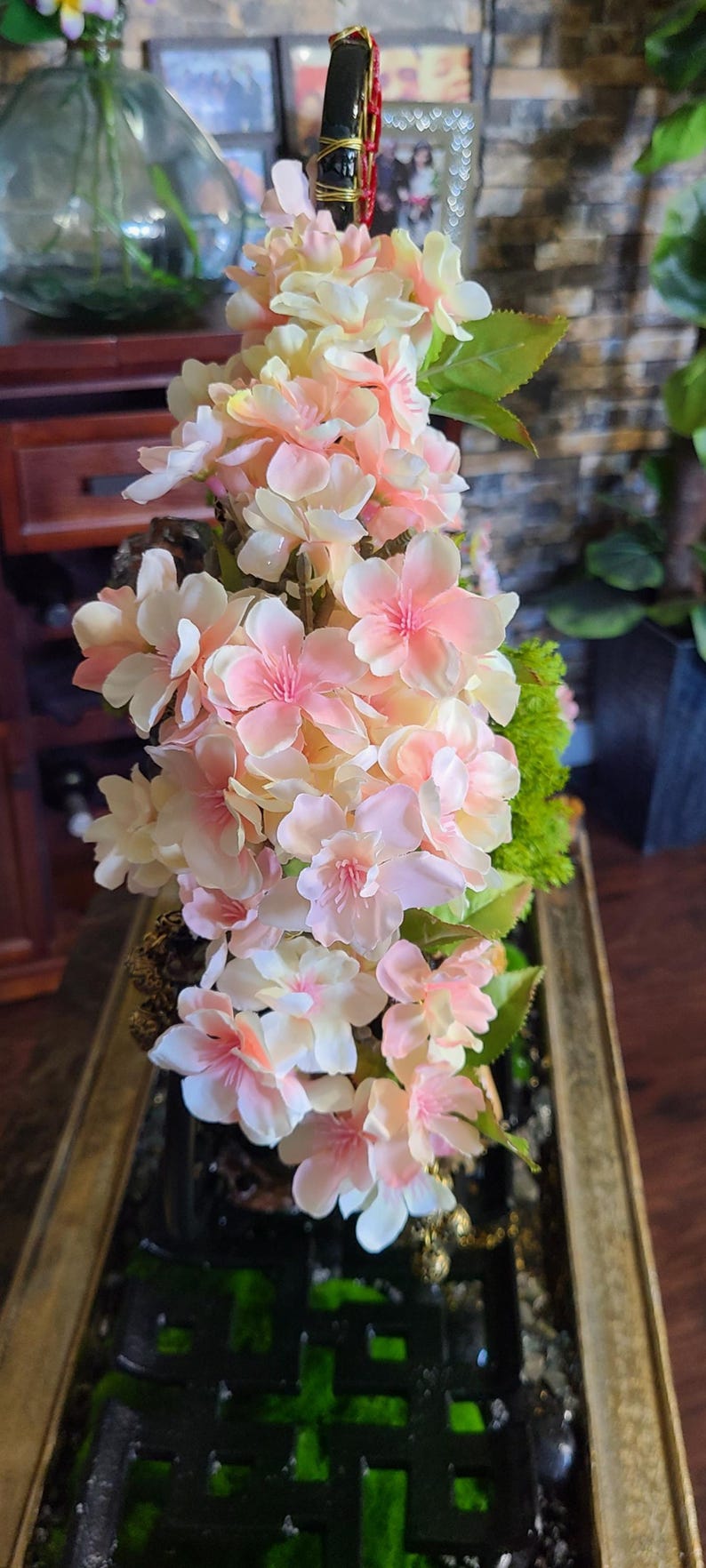 May include: A floral arrangement featuring delicate pink and white blossoms with green leaves. The flowers are arranged in a rectangular, gold-toned container with a dark green interior, creating a decorative display.