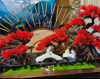 May include: A tabletop zen garden with three red bonsai trees, a white bridge, and two small figurines. The background features a wooden fan and a mountain scene.