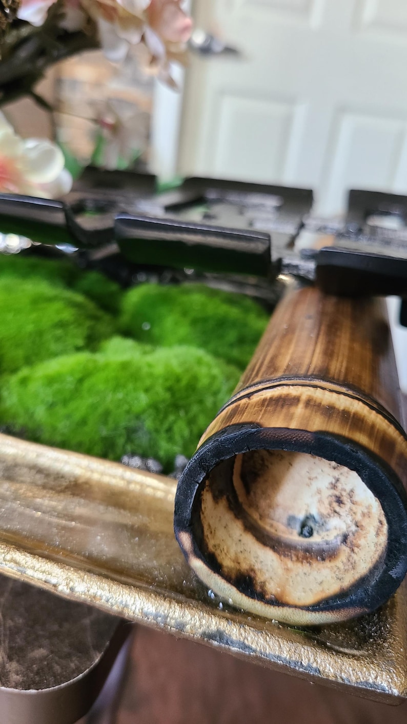 May include: Close-up of a bamboo tube with a charred rim, set against a backdrop of vibrant green moss. The bamboo has a natural wood grain and is partially inside a gold-colored tray. Decorative floral elements are visible in the background.
