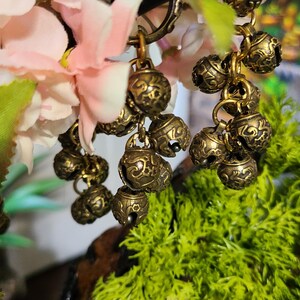 May include: Close-up of antique brass-colored bell charms hanging from a decorative branch. The bells have an ornate, embossed design and are clustered together. Pink and white flowers and green foliage provide a soft background.