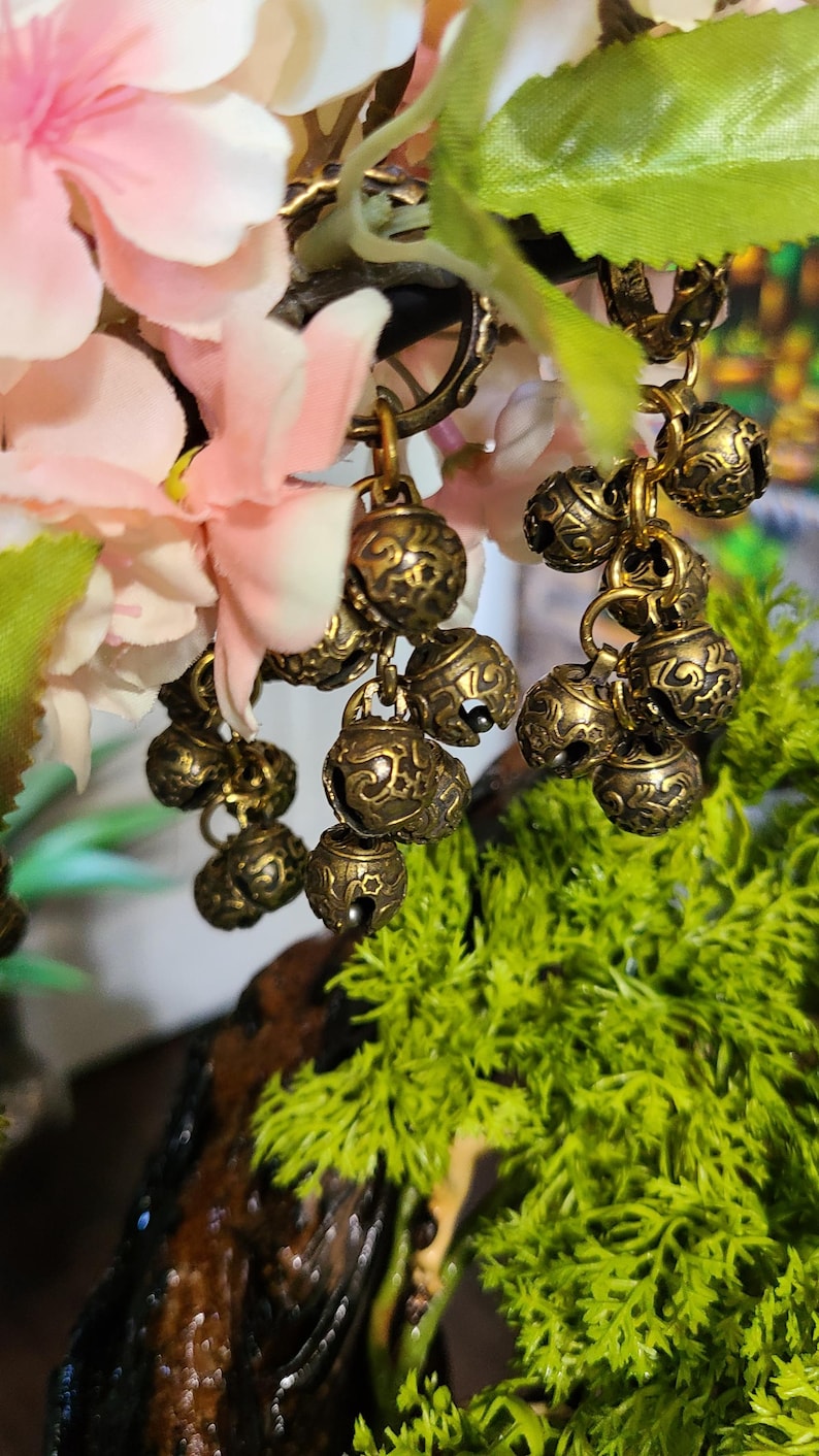May include: Close-up of antique brass-colored bell charms hanging from a decorative branch. The bells have an ornate, embossed design and are clustered together. Pink and white flowers and green foliage provide a soft background.