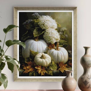 Könnte beinhalten: Ein gerahmter Kunstdruck, der ein Stillleben aus drei weißen Kürbissen zeigt, die zwischen Herbstblättern und weißen Hortensienblüten liegen. Die Szene hat einen dunklen, stimmungsvollen Hintergrund mit warmem, goldenem Licht auf den Texturen.