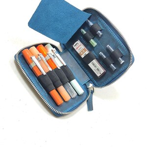 May include: A blue leather zippered case with multiple slots for pens and ink cartridges. The case is open and shows a variety of pens and cartridges, including a red and orange pen, a black pen, and a green pen.