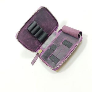 K&ouml;nnte beinhalten: Ge&ouml;ffnete lila Leder-Stiftemappe mit Rei&szlig;verschluss. Der Innenraum hat schwarze elastische Schlaufen und Taschen. Die H&uuml;lle ist ge&ouml;ffnet und gibt den Blick ins Innere frei. Auf wei&szlig;em Hintergrund.