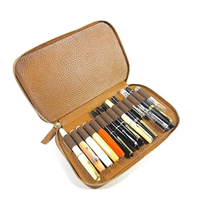 May include: A brown leather pen case, open to reveal a collection of fountain pens. The pens vary in color, including black, gold, white, and orange, with gold-colored nibs. The case has a zipper closure and a textured surface.