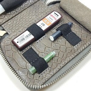 May include: A gray leather zippered pouch with multiple elastic loops for holding pens, pencils, and other writing instruments. The pouch is open and shows a variety of writing tools, including a green ink cartridge, a black mechanical pencil, and a brown mechanical pencil with the text "rotring tikky hi-polymer 0.5 2B 12" printed on it.