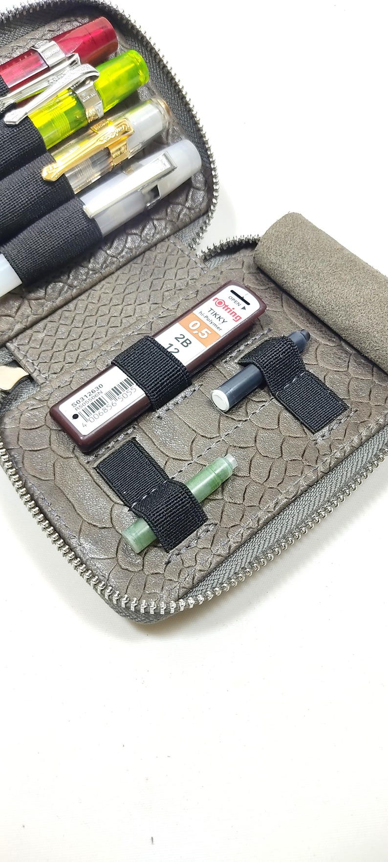 May include: A gray leather zippered pouch with multiple elastic loops for holding pens, pencils, and other writing instruments. The pouch is open and shows a variety of writing tools, including a green ink cartridge, a black mechanical pencil, and a brown mechanical pencil with the text "rotring tikky hi-polymer 0.5 2B 12" printed on it.