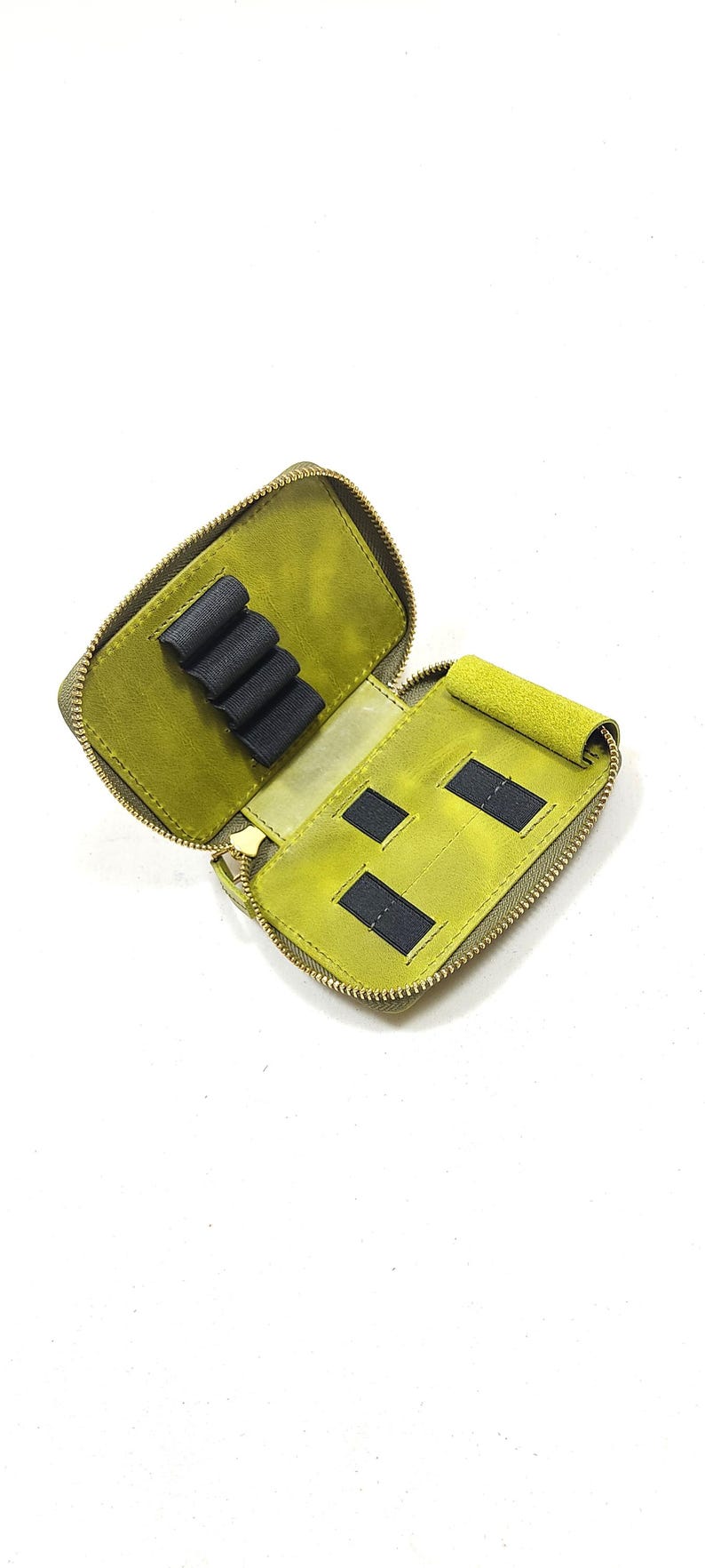 May include: Open, olive green leather pencil case with a gold zipper. The interior features black elastic loops and two black rectangular pockets. The case is designed to hold pens, pencils, or other small items.