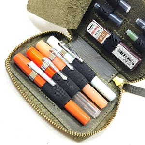 May include: A green leather zippered pouch with a flap, containing various writing instruments, including pens, pencils, and markers. The pouch has black elastic bands to hold the writing instruments in place.