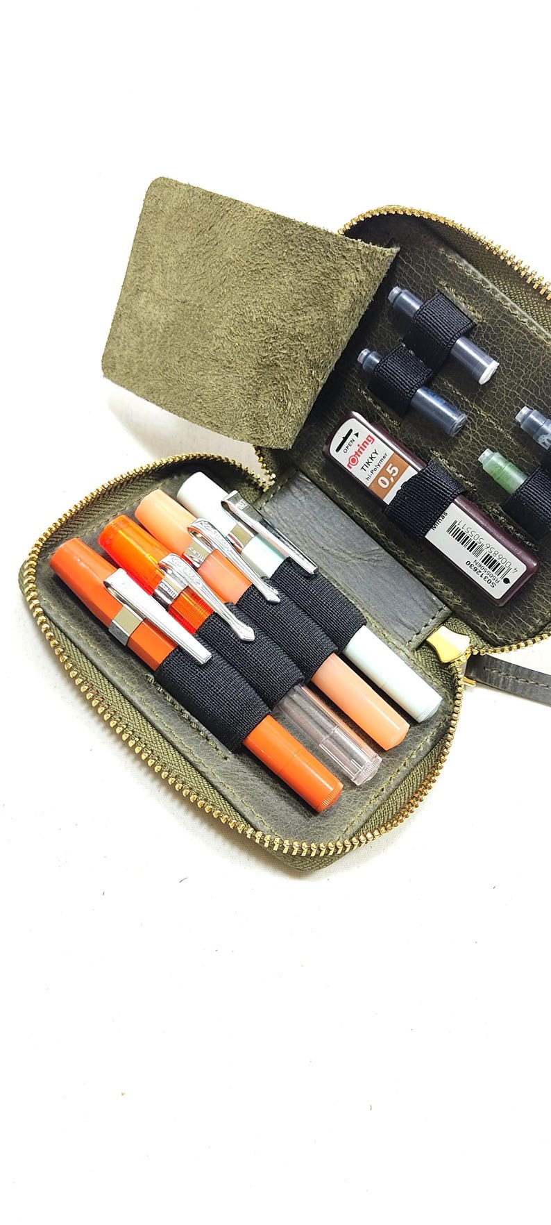 May include: A green leather zippered pouch with a flap, containing various writing instruments, including pens, pencils, and markers. The pouch has black elastic bands to hold the writing instruments in place.
