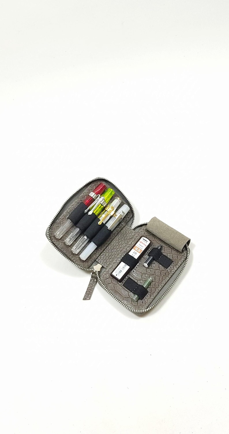 May include: A gray leather zippered pouch with a snakeskin pattern, open to reveal a variety of pens, pencils, and other writing implements.