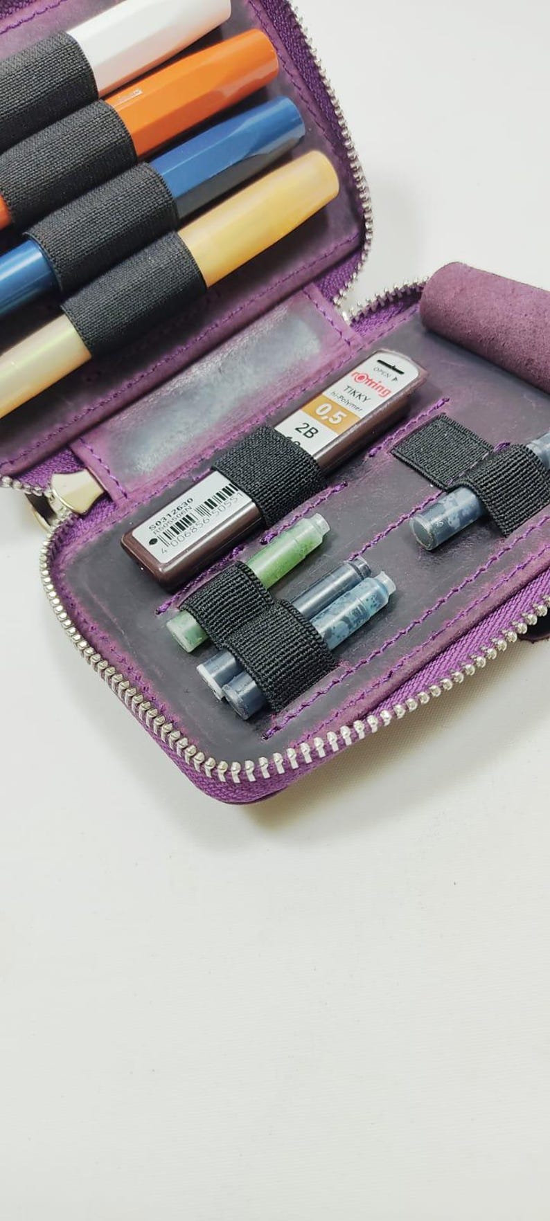 K&ouml;nnte beinhalten: Ein lila Leder-Federm&auml;ppchen mit Rei&szlig;verschluss, das mehrere farbige Stifte und Tintenpatronen enth&auml;lt. Das Etui hat schwarze Gummib&auml;nder zur Sicherung der Stifte und Patronen. Die Stifte sind wei&szlig;, orange, blau und gelb.