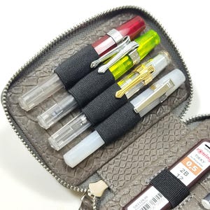 May include: A gray leather zippered pouch with a snakeskin pattern. The pouch is open and contains several pens, pencils, and a small brown rectangular object with a barcode and the text "S0312630 FRAGMENT".