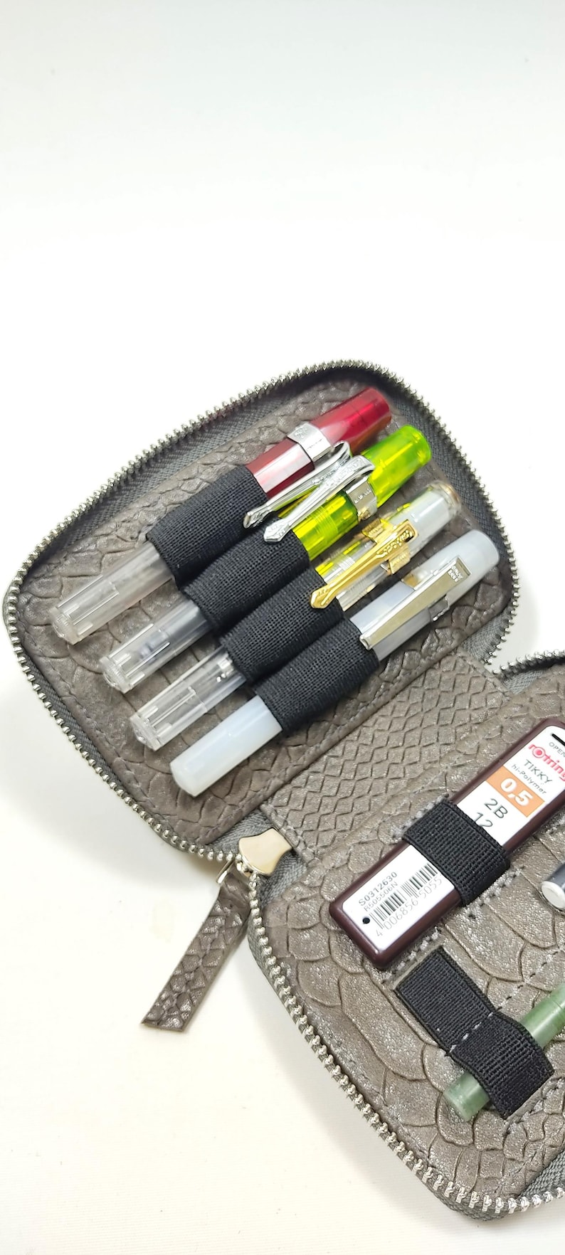 May include: A gray leather zippered pouch with a snakeskin pattern. The pouch is open and contains several pens, pencils, and a small brown rectangular object with a barcode and the text "S0312630 FRAGMENT".