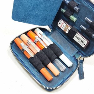 May include: A blue leather zippered pouch with a flap, containing several pens, pencils, and ink cartridges. The pouch has a zipper closure and a black elastic band to hold the writing instruments in place.