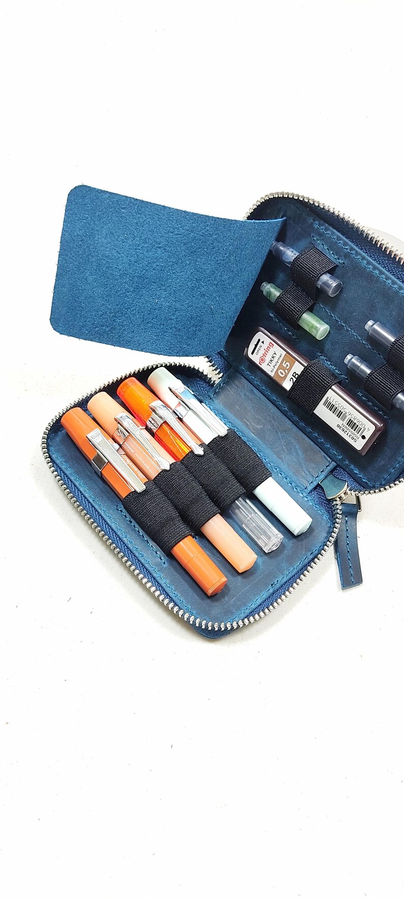 May include: A blue leather zippered pouch with a flap, containing several pens, pencils, and ink cartridges. The pouch has a zipper closure and a black elastic band to hold the writing instruments in place.