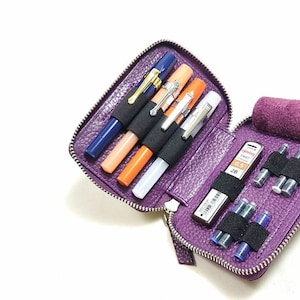 May include: Purple leather zippered case with multiple slots for pens, pencils, and ink cartridges. The case is open and shows a blue pen, two orange pens, a white pen, and a mechanical pencil with a 0.5 mm lead. There are also several ink cartridges in the case.