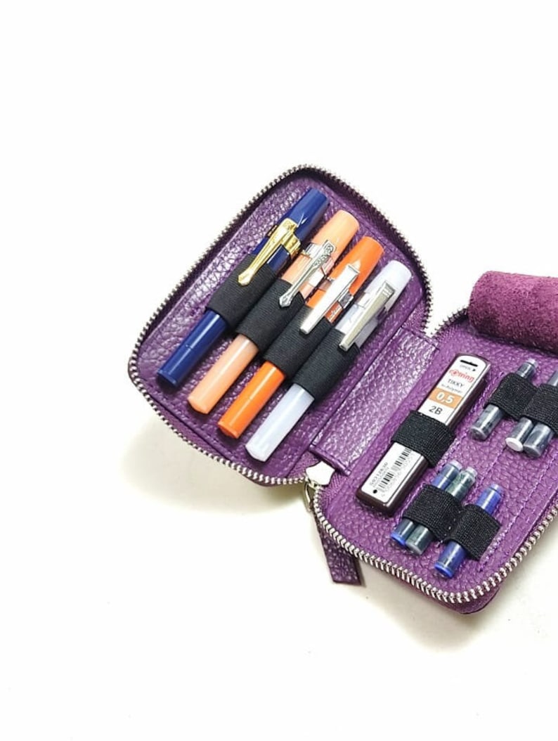 May include: Purple leather zippered case with multiple slots for pens, pencils, and ink cartridges. The case is open and shows a blue pen, two orange pens, a white pen, and a mechanical pencil with a 0.5 mm lead. There are also several ink cartridges in the case.