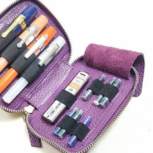 May include: Purple leather zippered pouch with multiple compartments for pens, pencils, and ink cartridges. The pouch is open and shows a black mechanical pencil, a silver mechanical pencil, and several blue ink cartridges.