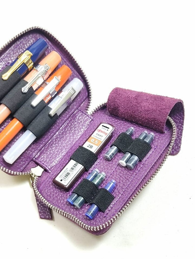 May include: Purple leather zippered pouch with multiple compartments for pens, pencils, and ink cartridges. The pouch is open and shows a black mechanical pencil, a silver mechanical pencil, and several blue ink cartridges.