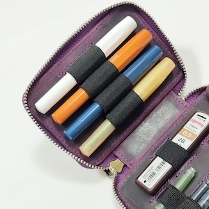 K&ouml;nnte beinhalten: Ein lila Stiftetui mit Rei&szlig;verschluss, gef&uuml;llt mit farbigen Stiften in Wei&szlig;, Orange, Blau und Gelb. Es enth&auml;lt auch Minen f&uuml;r Druckbleistifte. Die Stifte werden von einem schwarzen Gummiband gehalten.