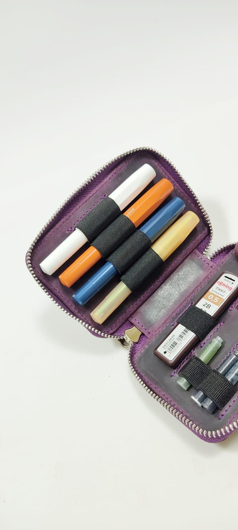 K&ouml;nnte beinhalten: Ein lila Stiftetui mit Rei&szlig;verschluss, gef&uuml;llt mit farbigen Stiften in Wei&szlig;, Orange, Blau und Gelb. Es enth&auml;lt auch Minen f&uuml;r Druckbleistifte. Die Stifte werden von einem schwarzen Gummiband gehalten.