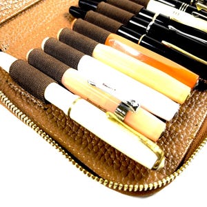 May include: A brown leather pen case with a gold zipper, filled with various fountain pens. The pens are in different colors, including black, white, orange, and cream, with gold and silver accents. The case is open, revealing the pens.